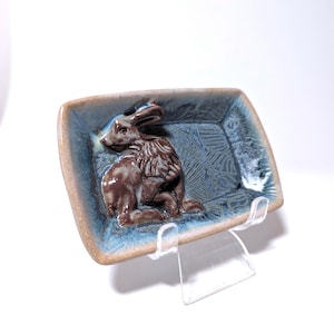 May include: A ceramic dish with a blue glaze and a brown rabbit sculpture in the center. The dish is rectangular and has a slightly raised edge. The rabbit is sitting on its haunches with its ears perked up.