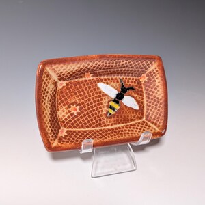 Puede incluir: Un plato rectangular de cer&aacute;mica con una superficie marr&oacute;n texturizada y una abeja decorativa. La abeja tiene alas blancas, cuerpo negro y rayas amarillas. El plato tiene un patr&oacute;n de rejilla y peque&ntilde;os detalles de estrellas.