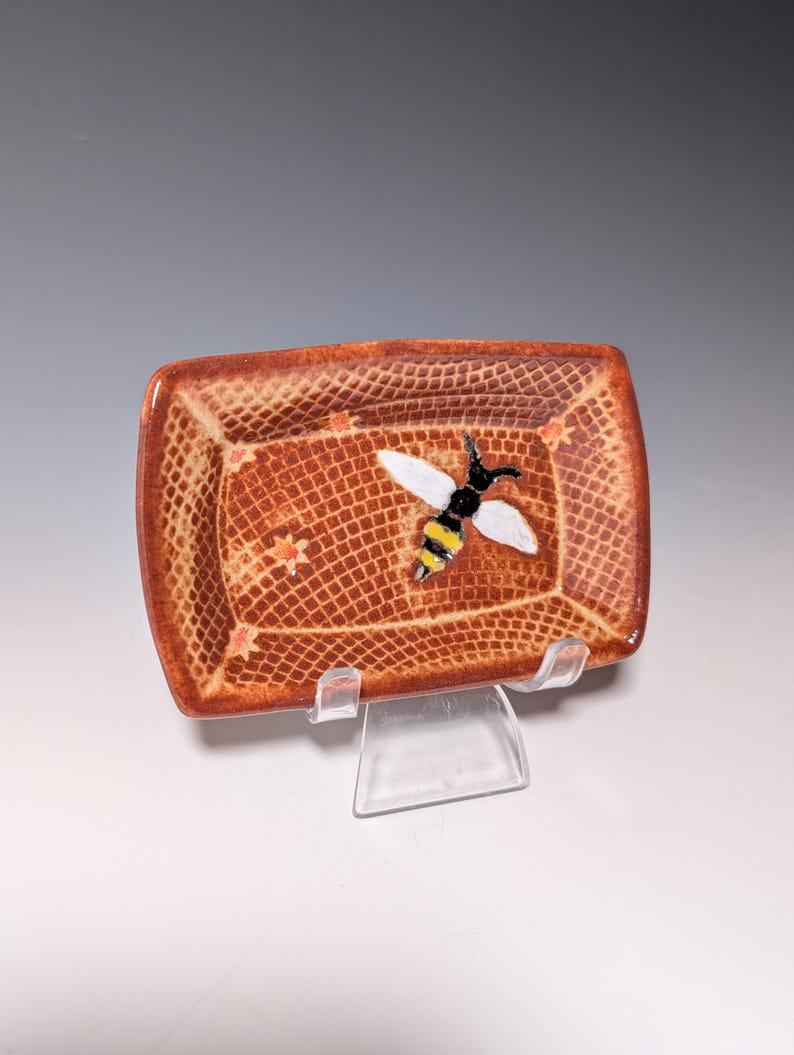 Puede incluir: Un plato rectangular de cer&aacute;mica con una superficie marr&oacute;n texturizada y una abeja decorativa. La abeja tiene alas blancas, cuerpo negro y rayas amarillas. El plato tiene un patr&oacute;n de rejilla y peque&ntilde;os detalles de estrellas.