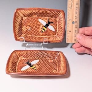 Puede incluir: Dos platos rectangulares de cer&aacute;mica marr&oacute;n con un patr&oacute;n de trama cruzada texturizada. Cada plato presenta una abeja pintada con alas blancas, cuerpo negro y rayas amarillas. Los platos miden aproximadamente 7.6 cm de largo.