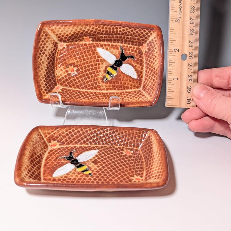 Puede incluir: Dos platos rectangulares de cer&aacute;mica marr&oacute;n con un patr&oacute;n de trama cruzada texturizada. Cada plato presenta una abeja pintada con alas blancas, cuerpo negro y rayas amarillas. Los platos miden aproximadamente 7.6 cm de largo.