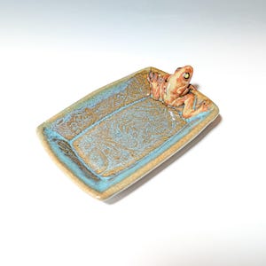 May include: A rectangular ceramic dish with a frog figurine. The dish has a turquoise and brown glaze with a floral pattern. The frog is orange and tan with green eyes. The dish is a decorative item.