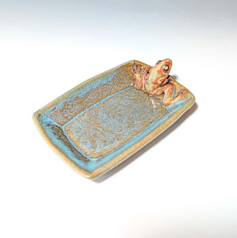 May include: A rectangular ceramic dish with a frog figurine. The dish has a turquoise and brown glaze with a floral pattern. The frog is orange and tan with green eyes. The dish is a decorative item.