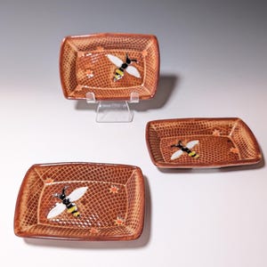 Puede incluir: Tres platos rectangulares de cer&aacute;mica con esquinas redondeadas. Cada plato es marr&oacute;n con un patr&oacute;n texturizado y una abeja pintada. Las abejas tienen alas blancas, cuerpos negros y rayas amarillas. Tambi&eacute;n hay peque&ntilde;os acentos de flores naranjas.