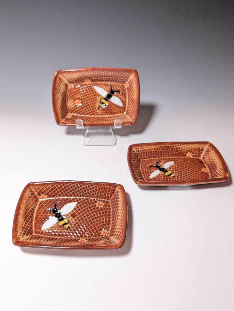 Puede incluir: Tres platos rectangulares de cer&aacute;mica con esquinas redondeadas. Cada plato es marr&oacute;n con un patr&oacute;n texturizado y una abeja pintada. Las abejas tienen alas blancas, cuerpos negros y rayas amarillas. Tambi&eacute;n hay peque&ntilde;os acentos de flores naranjas.