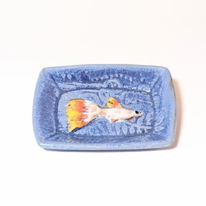 May include: A small, rectangular, blue ceramic dish with a textured surface. The dish features a detailed, hand-painted guppy fish with a yellow, orange, and white tail. The fish is centered in the dish, creating a decorative accent.
