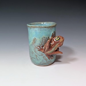 May include: A teal ceramic mug with a brown frog handle. The frog has a yellow eye and is climbing up the side of the mug.