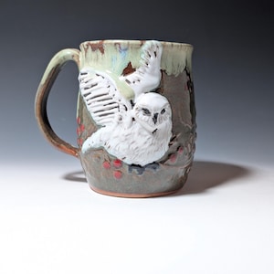 May include: A ceramic mug with a textured brown and green glaze featuring a raised, white sculpted owl with wings spread. The design includes small red berries on branches surrounding the owl, with a curved handle on the left side of the vessel.