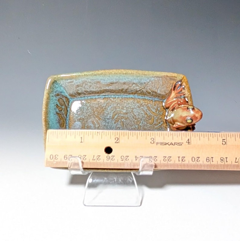May include: A rectangular ceramic dish with a fish-shaped handle. The dish is approximately 4 inches long and features a brown and teal glaze with a textured pattern. The fish handle is a light brown color.
