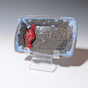 May include: A rectangular ceramic dish with a raised red cardinal figure on the left side. The dish has a blue rim and a textured gray interior. The dish is displayed on a clear plastic stand.