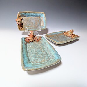 May include: Three ceramic serving trays with a frog design. The trays are rectangular with a turquoise and brown glaze. Each tray has a frog figurine attached to it. The trays are displayed on a white surface.