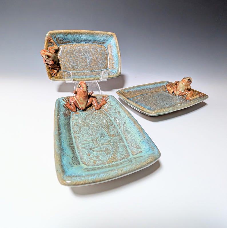 May include: Three ceramic serving trays with a frog design. The trays are rectangular with a turquoise and brown glaze. Each tray has a frog figurine attached to it. The trays are displayed on a white surface.