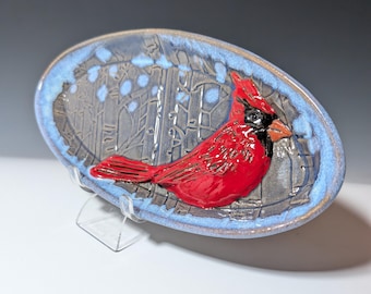 Puede incluir: Plato ovalado de cerámica con un relieve de un cardenal rojo. El plato tiene un borde azul y un interior gris con un diseño de escena invernal. El cardenal tiene detalles en negro y un pico naranja.