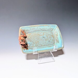 May include: A rectangular ceramic dish with a turquoise glaze and a brown frog figurine on the side. The dish has a textured surface with etched designs of mushrooms and plants. The frog has green eyes.