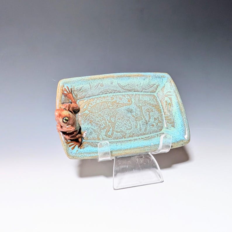 May include: A rectangular ceramic dish with a turquoise glaze and a brown frog figurine on the side. The dish has a textured surface with etched designs of mushrooms and plants. The frog has green eyes.