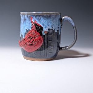 May include: A ceramic mug featuring a raised, sculpted red cardinal bird with black facial markings. The mug has a dark gray base with vertical texture and a light blue glaze dripping from the rim. It includes a curved handle on the right side.