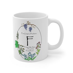 May include: White ceramic mug with a floral design featuring the letter F and the names of various flowers, including Forget Me Nots, Feverfew, and Foamflower. The text "Fox Grapes, Fleabane" is also included.