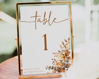 May include: Gold framed table number card with the number "1" and the text "table" in brown script. The card has a watercolor floral design in shades of brown, yellow, and blue. The text "TAYLOR + JUSTIN NOVEMBER 9TH 2024" is printed at the bottom of the card.