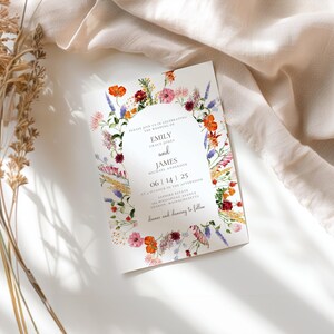May include: A wedding invitation with a floral wreath design in shades of pink, red, orange, yellow, and blue. The text reads "Emily and James" with the date "06 | 14 | 25".