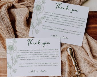 May include: Two white thank you cards with green floral accents and handwritten text. The cards are on a wooden surface with a beige cloth. A gold wax seal and stamps are also present.