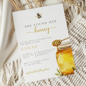 K&ouml;nnte beinhalten: Eine wei&szlig;e Brautduscheinladung mit goldener Schrift, die "She Found Her Honey" lautet. Die Einladung zeigt eine Aquarell-Illustration eines Honigglases mit einem Honigl&ouml;ffel und einer Biene.