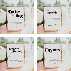 May include: Four gold-framed signs with white backgrounds and black text. The signs read "Date Night Jar", "Let Love Sparkle", "S'mores Station", and "Favors".