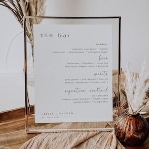 May include: A clear framed sign with the text "the bar" at the top. The sign lists wine, beer, spirits, and signature cocktail options. The names Olivia and Nathan are at the bottom, along with the date 10.18.2028.