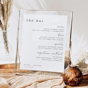 May include: A clear framed sign with the words "the bar" at the top. The sign lists wine, beer, spirits, and signature cocktail options. The names Olivia and Nathan are at the bottom, with the date 10.16.2025.