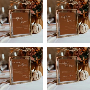 May include: Four gold-framed signs with brown paper inserts and white calligraphy. The signs read "open bar", "capture the love", "audio guestbook", and "bubble send off".