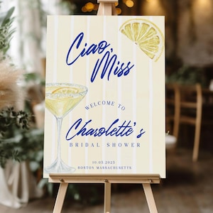 May include: A yellow and white striped sign with the text "Ciao, Miss" in blue cursive lettering. The sign also features a watercolor illustration of a margarita glass and a lemon slice. The text "Welcome to Charlotte's Bridal Shower" is in blue cursive lettering. The date "10.03.2023" and the location "Boston Massachusetts" are also included on the sign.