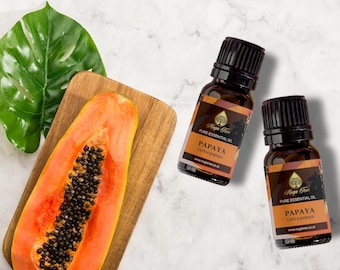 Könnte beinhalten: Zwei bernsteinfarbene Glasflaschen mit Papaya-Ätherischem Öl mit schwarzen Deckeln und goldenen Etiketten. Die Etiketten lauten "Kuga Free Pure Essential Oil Papaya Carica papaya" und enthalten eine Website-Adresse. Eine Hälfte einer Papaya-Frucht liegt auf einem Holzbrett.