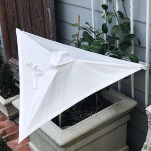 May include: A white square patio umbrella canopy featuring a central vent and a fabric tie closure. The canopy is made of solid white material and is positioned over a green plant in a square stone planter, showing a clean and simple design.