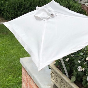 May include: A white square patio umbrella, open and angled, providing shade. The umbrella is made of a bright white fabric and is set up over a planter box filled with white flowers. The background includes green grass and a brick wall.