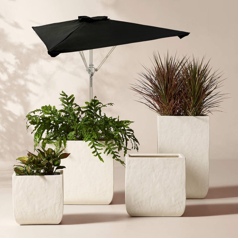 May include: A collection of four off-white square planters with a textured, matte finish. Two planters contain green foliage, while two are empty. A black patio umbrella with a silver metal pole stands behind the planters.