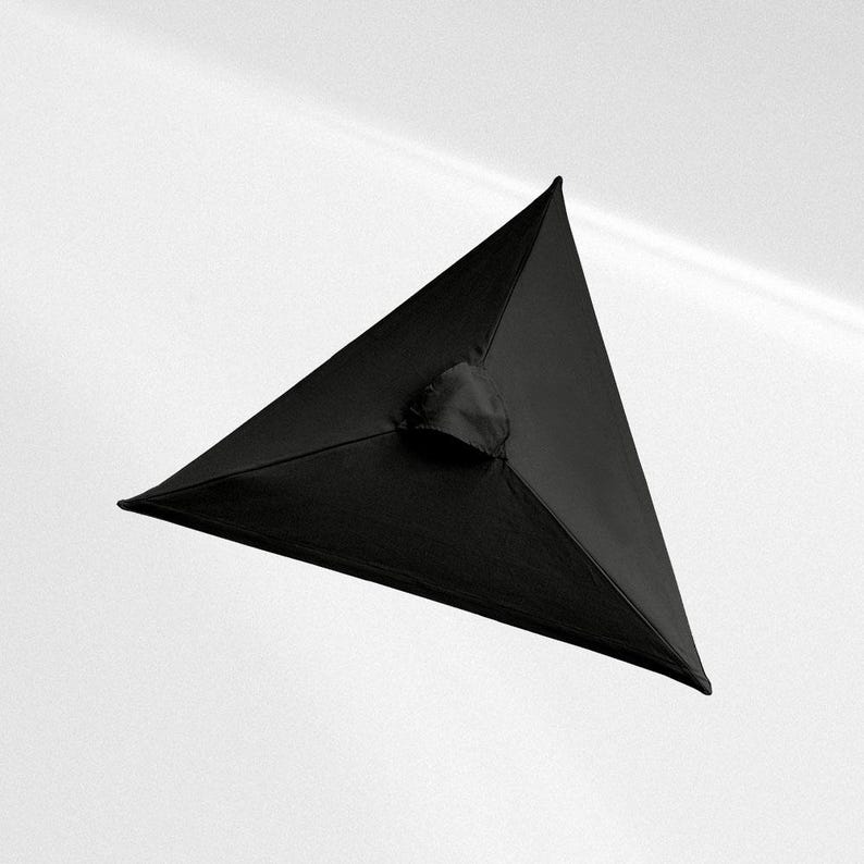 May include: A black, three-dimensional triangular pyramid object with smooth, matte fabric surfaces. The structure features sharp, defined edges meeting at a central apex, with a small, circular, gathered fabric opening located at the center.