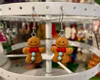 May include: A pair of gingerbread man earrings with red bows and white icing details. The earrings are hanging on a white earring display stand.