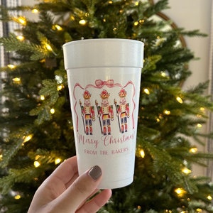 May include: White disposable cup with a pink ribbon and three nutcracker illustrations. The cup says "Merry Christmas from the Bakers".