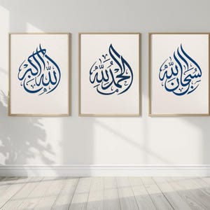 Puede incluir: Tres impresiones artísticas enmarcadas con caligrafía árabe en un interior moderno. Cada impresión presenta un diseño caligráfico diferente en azul oscuro sobre un fondo crema, enmarcado en madera clara. Las impresiones se exhiben en una pared blanca.