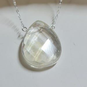 May include: A silver necklace featuring a faceted, teardrop-shaped clear crystal pendant. The pendant hangs from a delicate silver chain. The crystal has a multifaceted design, reflecting light. The necklace is displayed on a white surface.