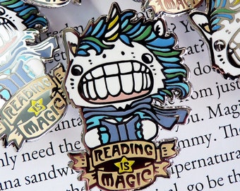 May include: A silver enamel pin with a cartoon unicorn holding a book. The unicorn has a blue and green mane and tail. The pin says "Reading is Magic" in a banner.