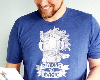May include: A man wearing a blue t-shirt with a white unicorn graphic and the text "Reading is Magic". He is holding an open book with illustrations.