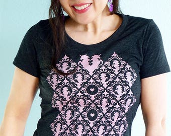 May include: A dark grey t-shirt with a pink and black pattern of stylized unicorns and hearts. The pattern is a repeating design and covers the entire front of the shirt.