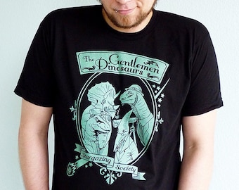 May include: A black t-shirt with a green graphic of dinosaurs wearing top hats and holding glasses. The text on the shirt reads "The Gentlemen Dinosaurs Stargazing Society."