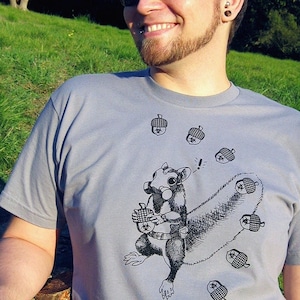 May include: A gray t-shirt with a black and white illustration of a squirrel wearing a gas mask and holding an acorn. The squirrel is surrounded by acorns. The text "Shroom Queens" is printed on the shirt.