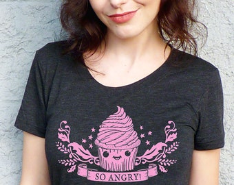 May include: Dark gray v-neck t-shirt featuring a pink cupcake graphic with the text "SO ANGRY!". The cupcake has a smiling face and is surrounded by stars and decorative elements. The shirt is made of a soft material.