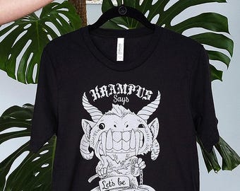 May include: Black t-shirt featuring a white illustration of Krampus with the text "Krampus Says" above the image and "Let's be Naughty" below. The shirt is hanging on a black hanger.