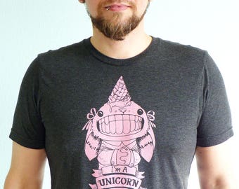 May include: A dark grey t-shirt with a pink cartoon creature wearing a pink ice cream cone hat. The creature has a large smile and the text "I'm a UNICORN Now" is printed below it.