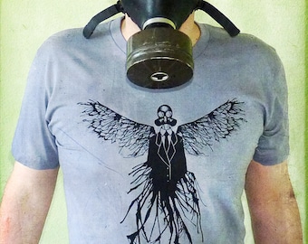 May include: A person wearing a light blue t-shirt with a black and white graphic of a winged figure wearing a gas mask and a suit. The figure has roots or vines extending from its body. The person is also wearing a black gas mask.