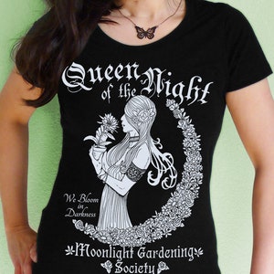 May include: Black t-shirt with a white graphic design featuring a woman holding a flower and the text "Queen of the Night", "We Bloom in Darkness", and "Moonlight Gardening Society".
