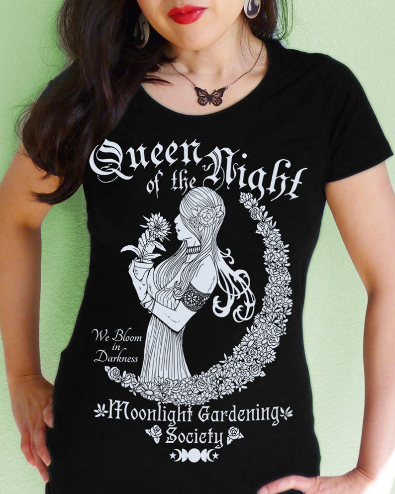 May include: Black t-shirt with a white graphic design featuring a woman holding a flower and the text "Queen of the Night", "We Bloom in Darkness", and "Moonlight Gardening Society".